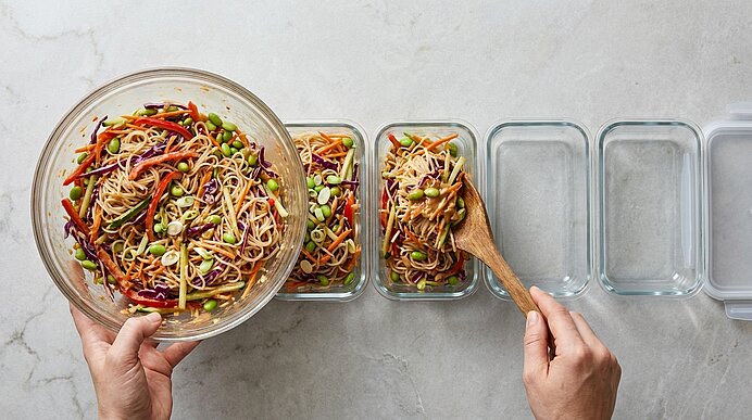 Hände portionieren aus einer großen Glasschüssel bunten Nudel‑Gemüse‑Salat (Karotten, Rotkohl, Edamame, Sesam) in mehrere rechteckige Glas‑Mealprep‑Behälter.