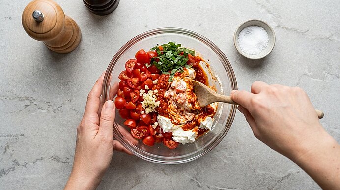 Zwei Hände rühren in einer Glasschüssel halbierte Cherrytomaten, Joghurt, gehackten Knoblauch, Kräuter und rote Gewürzpaste mit einem Holzlöffel zu einem Dip.