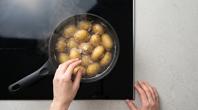 Draufsicht: Zwei Hände halten einen Topf mit kleinen Kartoffeln, die in kochendem Wasser auf dem Herd stehen; Dampf steigt auf.