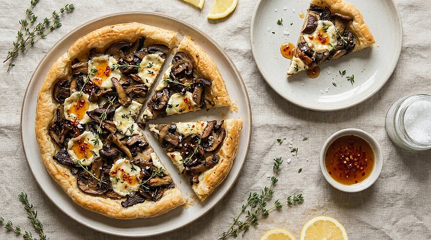 Herzhafte Pilztarte auf einem Teller: knuspriger Rand, geschmolzener Käse, gebratene Pilze, Kräuter und Chiliflocken; ein Stück auf einem kleinen Teller, daneben Zitronenscheiben und eine Schale Chilisoße.