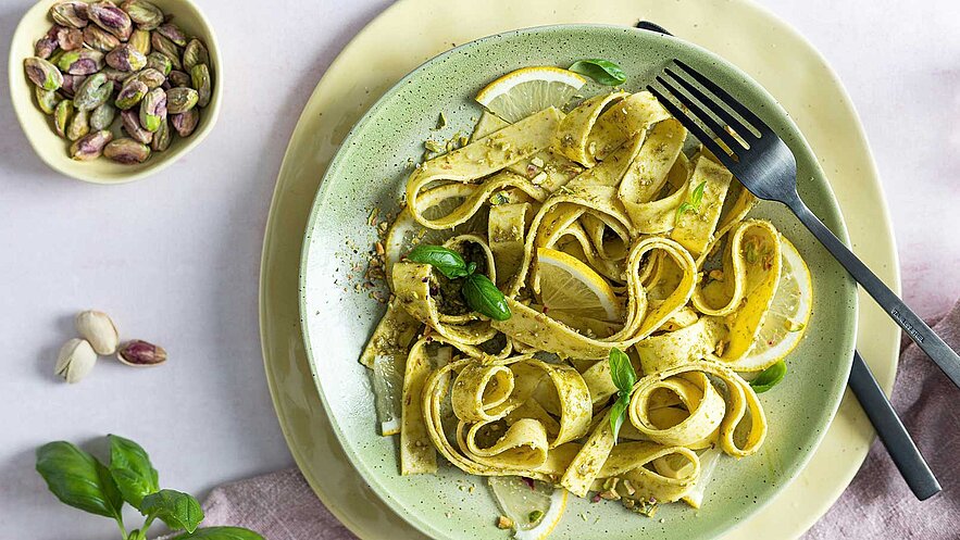 Ein Teller mit Bandnudeln in einer grünen Schale, garniert mit frischen Basilikumblättern und Zitronenscheiben. Die Nudeln sind mit einer Pesto-Sauce bestreut. Daneben eine kleine Schale mit Pistazien. Der Hintergrund ist schlicht und hell gehalten.