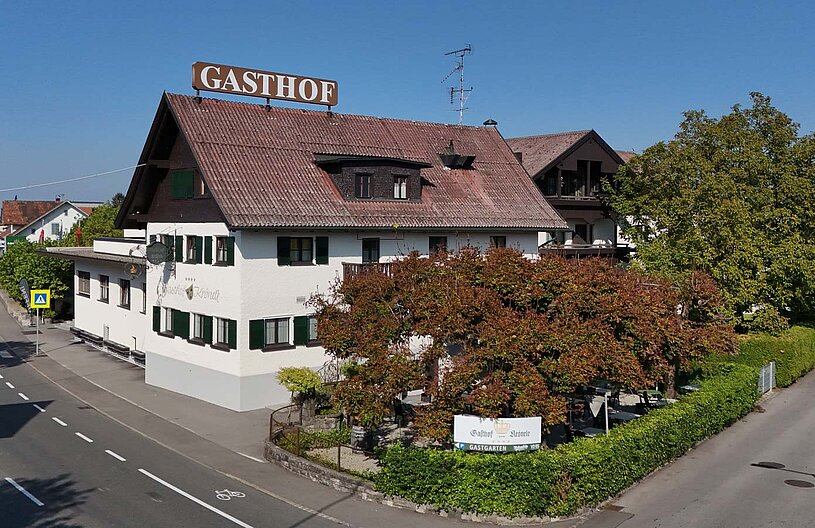 Ein Gasthof mit dem Schild „Gasthof“ auf dem Dach. Das Gebäude hat weiße Wände und grüne Fenster, umgeben von Bäumen und Sträuchern. Im Vordergrund ist die Straße zu sehen, die am Gasthof vorbeiführt. Der Himmel ist klar und blau.