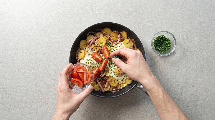 Zwei Hände belegen eine Pfanne mit Bratkartoffeln und Spiegelei mit frischen Tomatenscheiben; daneben eine kleine Schale mit Schnittlauch.