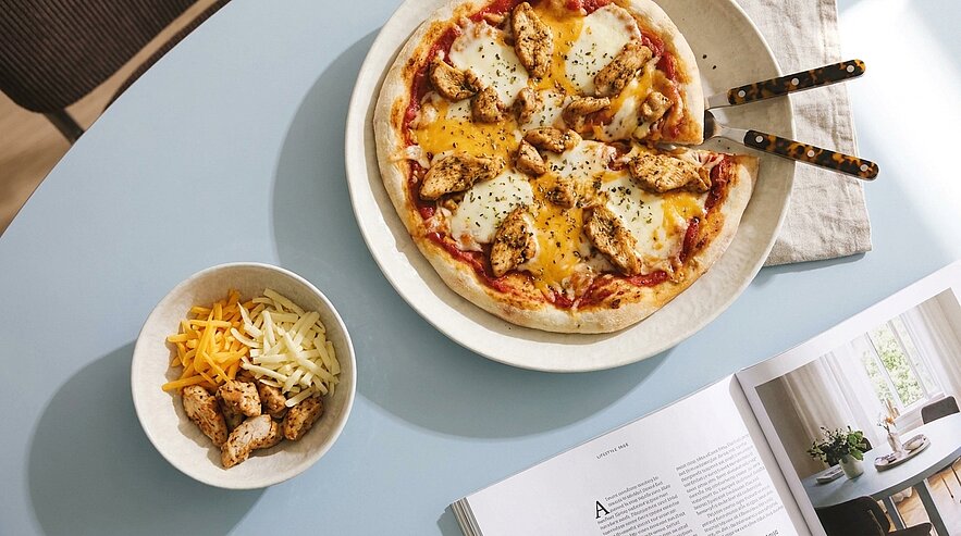 Pizza auf Teller mit geschmolzenem Käse, gebratenen Hähnchenstücken und Kräutern; daneben eine kleine Schale mit geriebenem Käse und weiteren Hähnchenstücken.