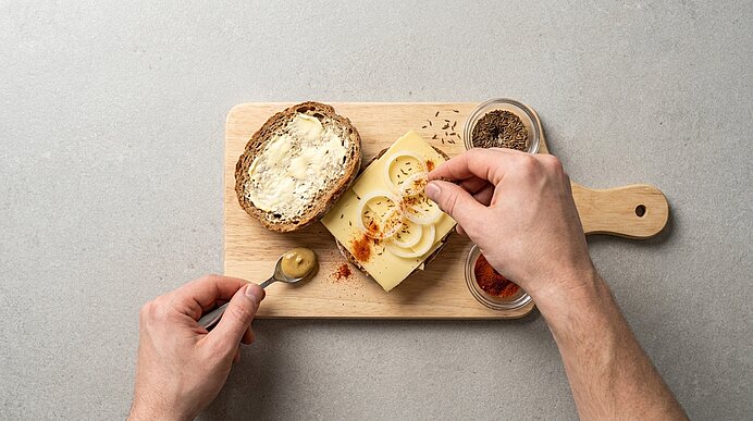 Hände belegen auf einem Schneidebrett eine Brotscheibe mit Käse und Gewürzgurken; daneben eine gebutterte Scheibe, Senf und Gewürze.