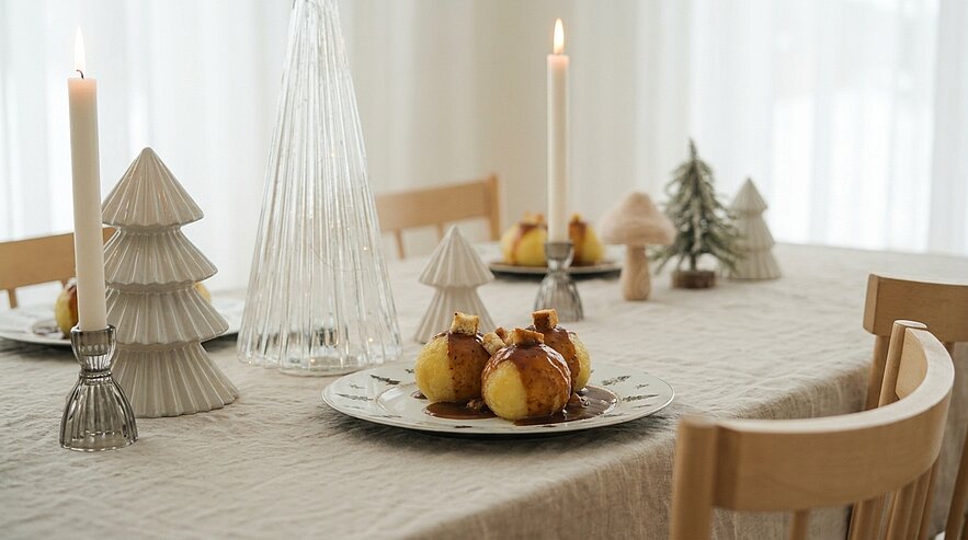 Festlich gedeckter Esstisch mit Teller, darauf drei Bratäpfel, brennenden Kerzen und kleinen Tannenbaum-Dekoren auf hellem Tischtuch.