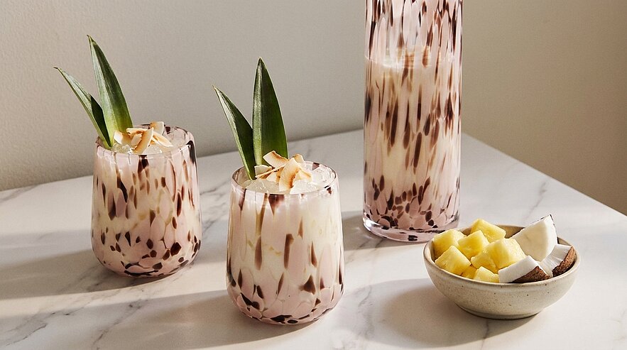 Zwei kurze Gläser und eine hohe Karaffe mit marmoriertem, cremigem Getränk; die Gläser sind mit Ananasblättern und Kokosflocken garniert. Daneben eine Schale mit Ananas- und Kokosstücken.
