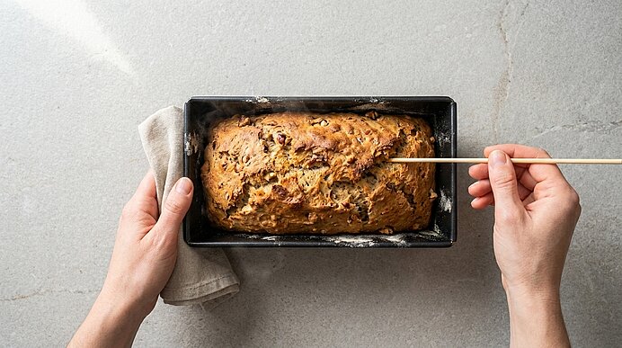 Hände halten eine Kastenform mit frisch gebackenem Brot; eine Hand mit Tuch, die andere führt einen Holzspieß in das Brot zur Garprobe.