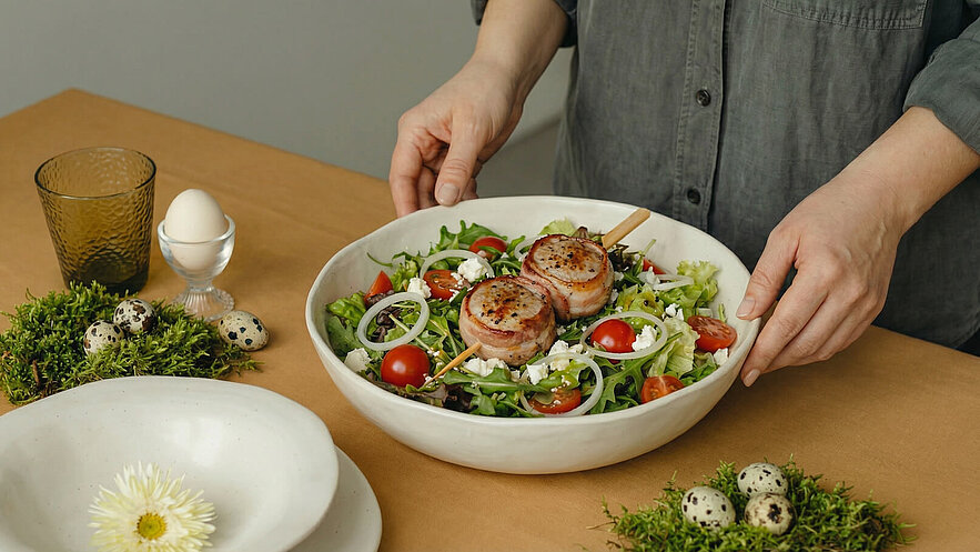 Eine Person präsentiert eine große Schüssel mit gemischtem Salat. Der Salat enthält grüne Blätter, Kirschtomaten, Zwiebelringe und Feta. Auf dem Salat liegen zwei mit Speck umwickelte Fleischscheiben. Im Hintergrund sind ein Glas, ein Ei und einige Wachteleier zu sehen.