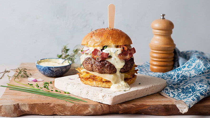 Ein üppiger Burger auf einem Holzbrett, gefüllt mit einem saftigen Patty, geschmolzenem Käse, Speck und frischen Zutaten. Eine kleine Schüssel mit Sauce befindet sich neben dem Burger. Dekorative Kräuter und Frühlingszwiebeln liegen auf dem Brett.