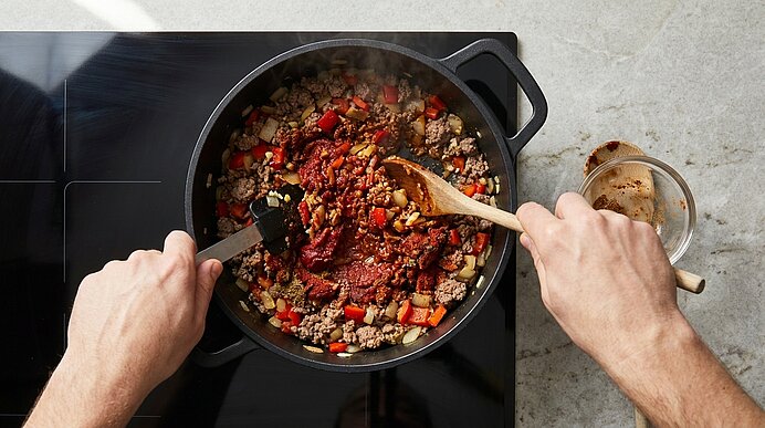 Zwei Hände verrühren angebratenes Hackfleisch, Zwiebeln, Paprika, Tomatenmark und Gewürze in einem schwarzen Topf.