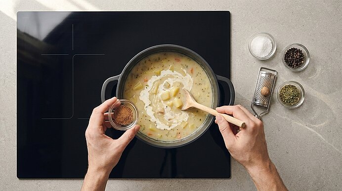 Von oben: Hände rühren in einem Topf cremige Kartoffelsuppe mit Holzlöffel; Sahne und Gewürze werden ergänzt, kleine Schälchen mit Salz, Pfeffer und Kräutern daneben.