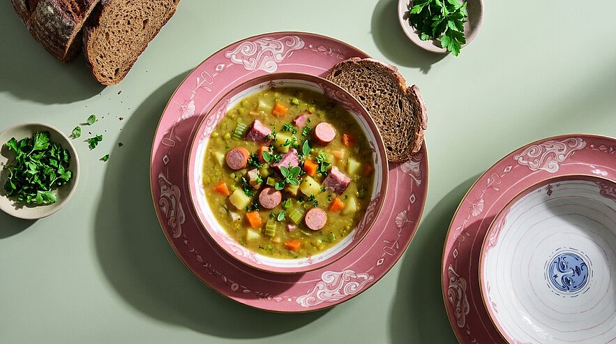 Schüssel mit herzhafter Gemüsesuppe (Kartoffeln, Karotten, Sellerie), kleinen Wurstscheiben und frischer Petersilie, serviert mit zwei Scheiben dunklem Brot.