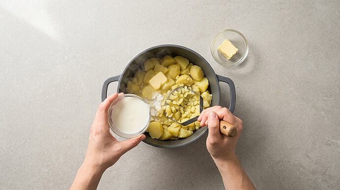 Hände stampfen gekochte Kartoffeln in einem Topf und fügen Milch sowie Butter hinzu, um Kartoffelpüree zuzubereiten.