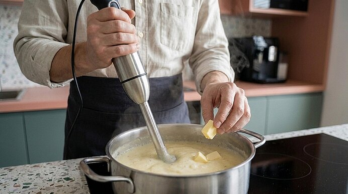 Person rührt mit Stabmixer in einem Topf und gibt Butter hinzu.