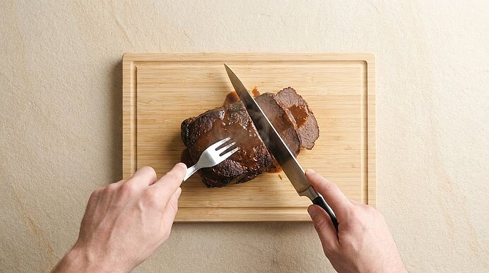 Zwei Hände schneiden ein gebratenes Stück Fleisch auf einem Holz-Schneidebrett mit Messer und Fleischgabel.