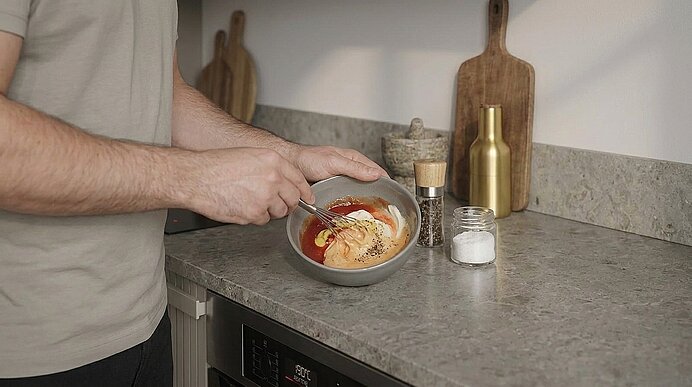Person rührt mit einem Löffel eine Sauce in einer Schüssel auf der Küchenarbeitsplatte; Salz- und Pfefferstreuer stehen daneben.