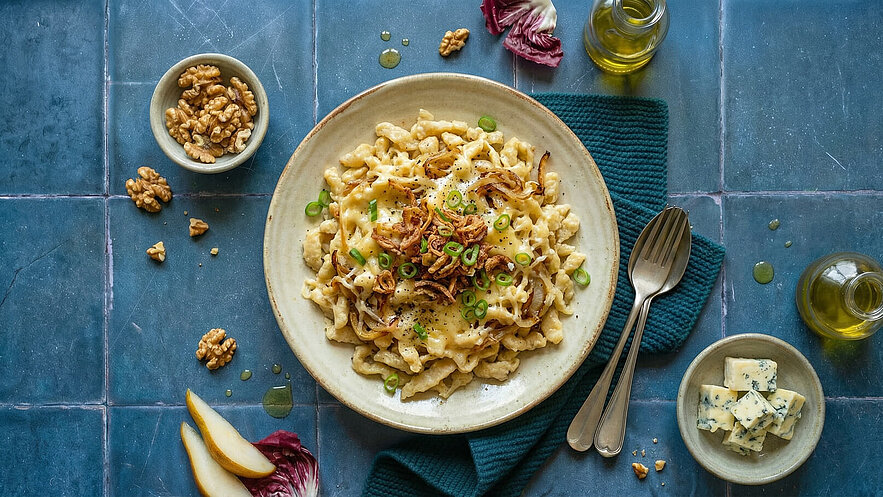 Ein Teller mit cremiger Pasta, garniert mit frittierten Zwiebeln und Frühlingszwiebeln. Daneben liegt eine Schale mit Walnüssen und eine weitere mit Blauschimmelkäsewürfeln. Das Ganze ist auf einer blauen Fliese angerichtet, umgeben von Olivenöl und einem Tuch.