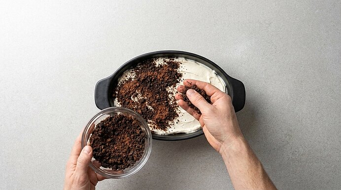 Zwei Hände streuen dunkle Schokoladenkekskrümel aus einer Schüssel auf die weiße Creme in einer schwarzen Form.