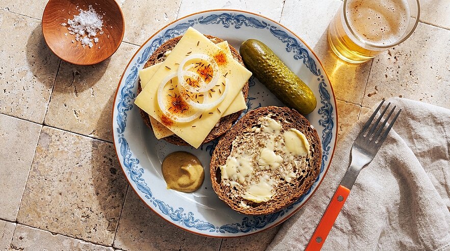 Teller mit zwei Scheiben Vollkornbrot, eine mit Butter, eine belegt mit Käse und Zwiebelringen; dazu Senf, Gewürzgurke, ein Glas Bier und Besteck auf einer Serviette.