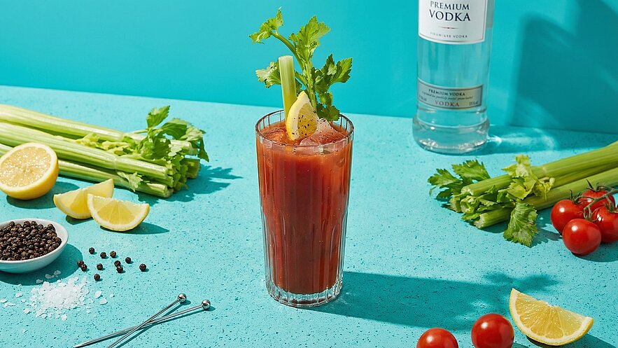Ein Glas Bloody Mary mit Eis, garniert mit Sellerie und einer Zitronenscheibe, steht auf einem türkisfarbenen Tisch. Um das Glas herum sind frische Zutaten wie Zitronen und Kirschtomaten verteilt, sowie Pfefferkörner und Salz.