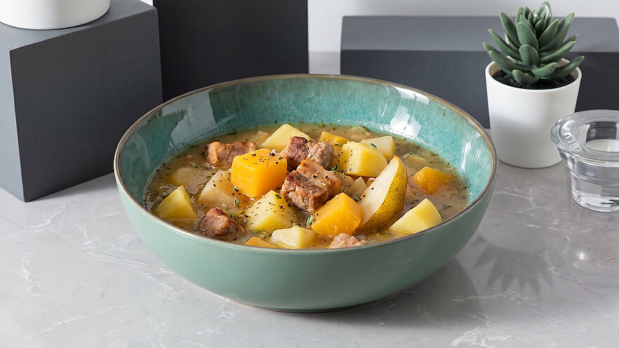 Eine grüne Schüssel enthält einen Eintopf mit Stückchen von Fleisch und verschiedenen Gemüsesorten wie Kürbis und Kartoffeln. Der Eintopf ist in einer klaren, brüheartigen Flüssigkeit serviert und hat eine appetitliche, reichhaltige Optik.