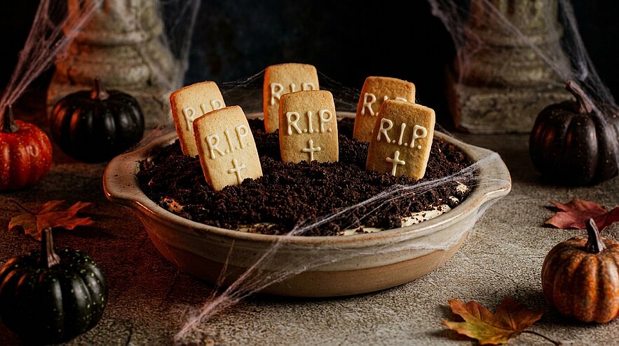 Halloween-Dessert: Schale mit krümeliger "Erde" und mehreren Keks-Grabsteinen mit "R.I.P."-Aufschrift, dekoriert mit kleinen Kürbissen.