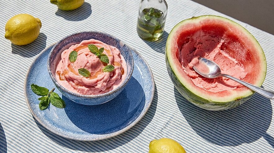 Schale mit rosa Wassermelonen-Sorbet, garniert mit Minzblättern; daneben eine ausgehöhlte halbe Wassermelone mit Löffel.