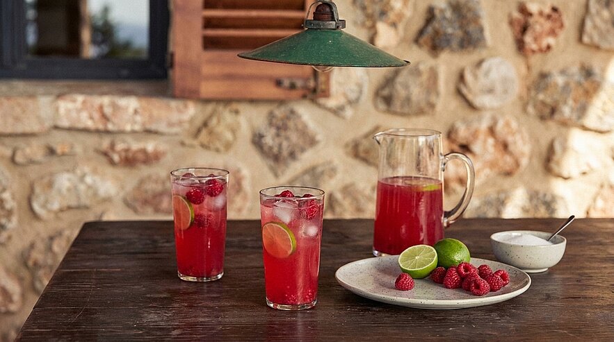 Auf einem Holztisch stehen drei Gläser mit einem roten Getränk, verziert mit Limettenscheiben und Himbeeren. Daneben befindet sich eine Karaffe mit dem gleichen Getränk sowie ein Teller mit weiteren Himbeeren und einer Limette. Der Hintergrund zeigt eine Steinmauer.