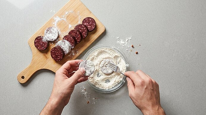 Zwei Hände panieren Salami-Scheiben: auf einem Holzbrett liegen mehrere Scheiben, daneben eine Glasschüssel mit Mehl; eine Hand hält eine Scheibe, die andere sticht mit einer Gabel ins Mehl.