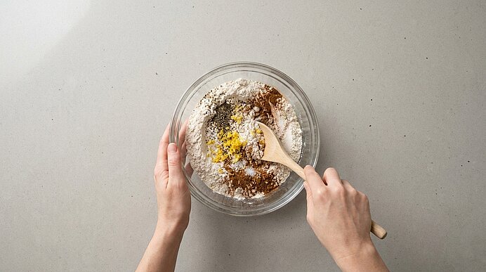 Zwei Hände rühren mit einem Holzlöffel Mehl und Gewürze in einer Glasschüssel auf einer hellen Arbeitsfläche.