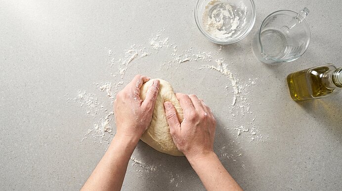 Zwei Hände kneten Pizzateig auf einer bemehlten Arbeitsfläche. Rechts stehen Schüssel, Messbecher und Olivenöl.