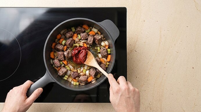 Von oben: Hände rühren in einem Kochtopf auf Cerankochfeld einen Eintopf aus gewürfeltem Rindfleisch, Karotten, Sellerie und Zwiebeln; mit Holzlöffel wird Tomatenmark hinzugefügt.