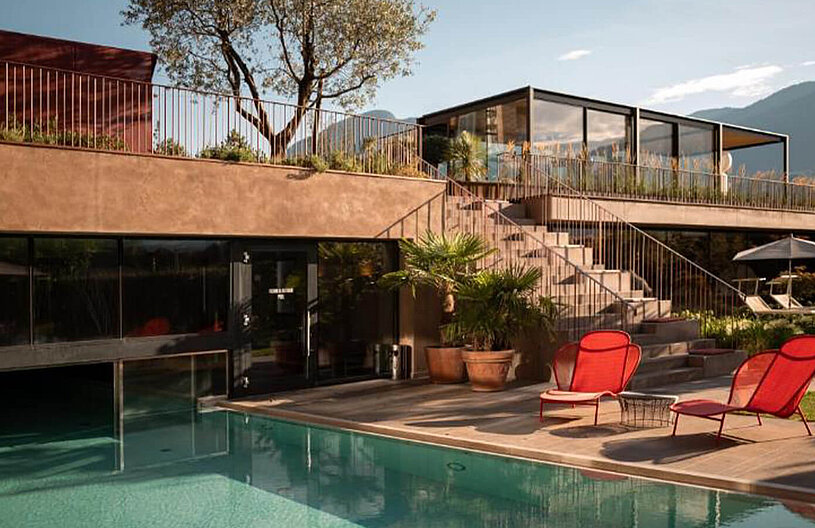 Modernes Haus mit großer Glasveranda und Terrasse, Außenpool im Vordergrund, zwei rote Liegestühle am Beckenrand, Treppe zur oberen Gartenterrasse und Bergkulisse.