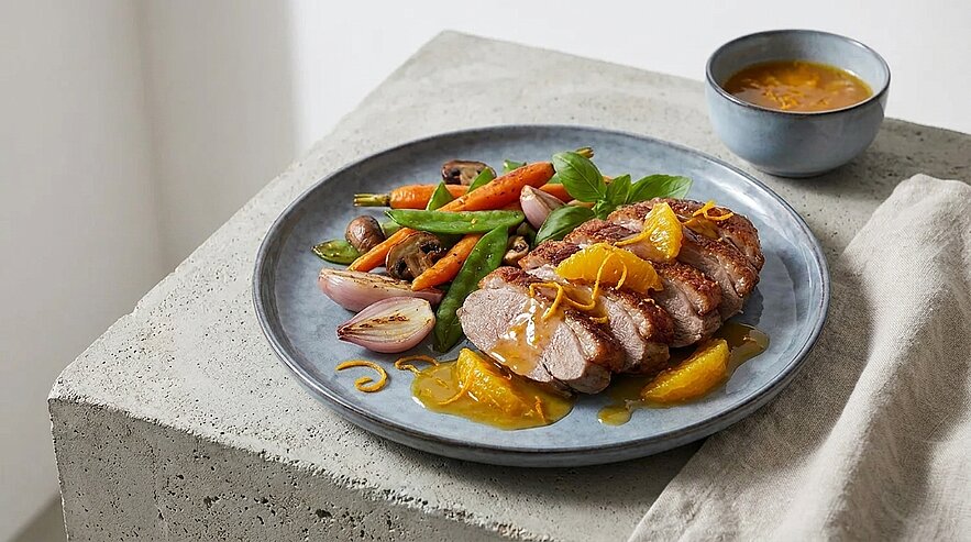 Gegartes, in Scheiben geschnittenes Entenbrustfilet mit Orangenscheiben und Sauce auf einem Teller, dazu gebratenes Gemüse (Karotten, Zuckerschoten, Schalotten) und eine kleine Schale mit zusätzlicher Sauce.