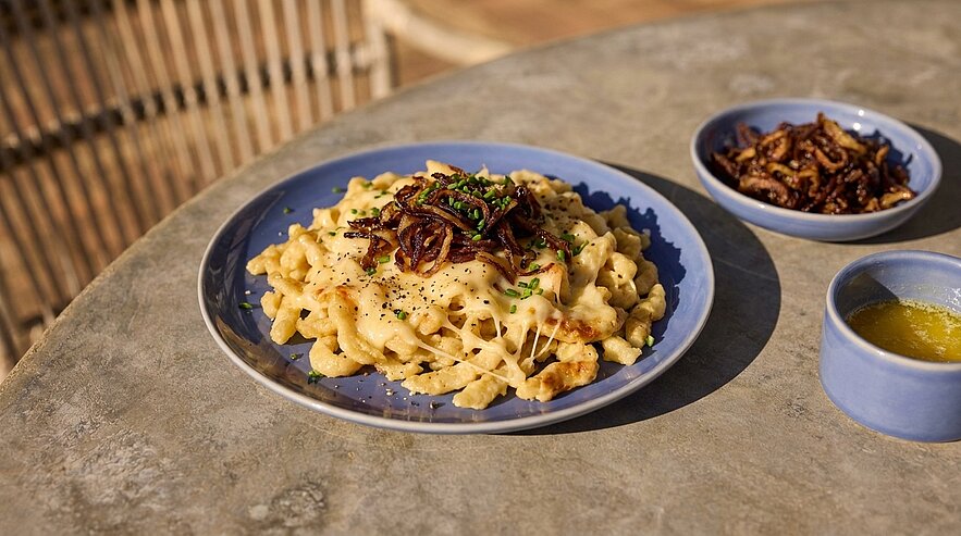 Teller mit cremigen Käsespätzle, garniert mit knusprigen Röstzwiebeln und Schnittlauch; daneben zwei kleine Schälchen mit zusätzlichen Röstzwiebeln und flüssiger Butter.