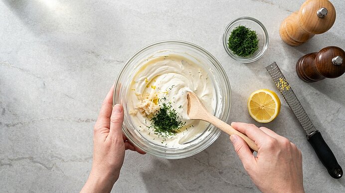 Zwei Hände rühren Frischkäsecreme mit Dill und Meerrettich in einer Glasschüssel mit einem Holzlöffel glatt.