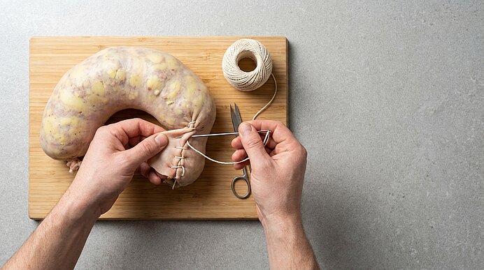 Hände binden mit Schnur und Schere die Hülle einer großen, rohen Ringwurst auf einem Holzbrett.