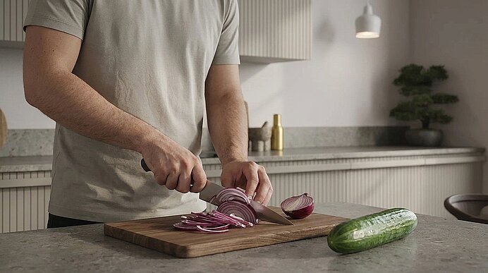 Person schneidet rote Zwiebel auf einem Holzbrett mit einem Messer; geschnittene Zwiebelringe und eine ganze Gurke liegen auf der Küchenarbeitsplatte.