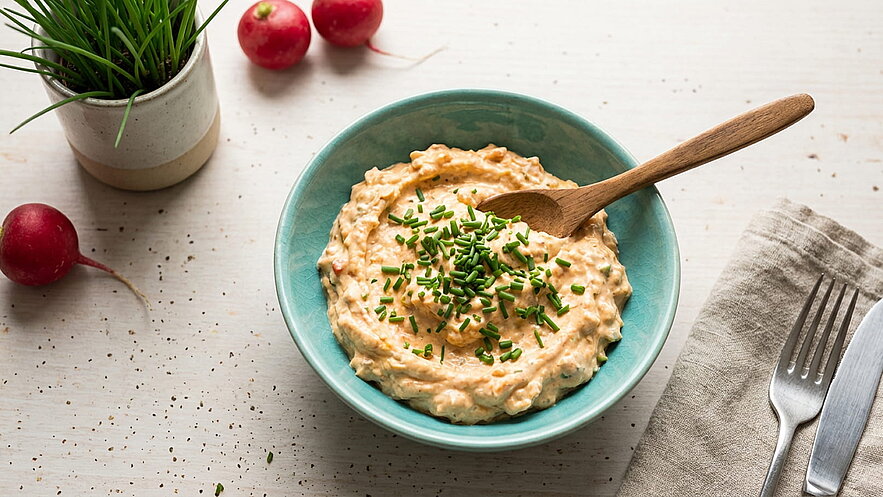 Eine Schüssel mit cremigem, orangenem Dip, garniert mit frischem Schnittlauch. Ein Holzlöffel liegt in der Schüssel. Im Hintergrund sind Radieschen und ein kleiner Blumentopf mit Gras zu sehen. Die gesamte Anordnung ist auf einem hellen Tisch platziert.
