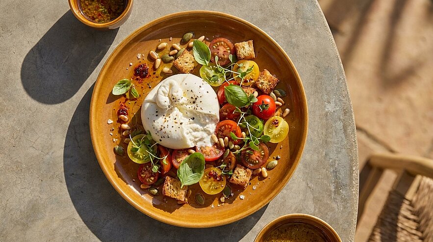 Teller mit cremiger Burrata in der Mitte, umgeben von bunten Kirschtomaten, frischem Basilikum, Croutons, Kürbis- und Pinienkernen sowie Olivenöl-Dressing.