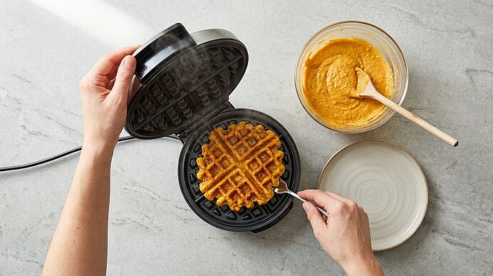 Geöffnete Waffeleisen mit goldbrauner Waffel; daneben Schüssel mit orangefarbenem Teig und Löffel sowie ein leerer Teller. Hände öffnen das Gerät und stechen die Waffel mit einer Gabel.