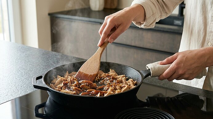Hände rühren mit Holzspatel gebratene Pilze in einer gusseisernen Pfanne auf dem Herd.