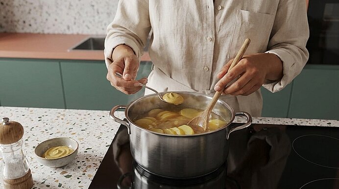 Person rührt mit Löffel und Holzlöffel in einem großen Kochtopf auf dem Herd, darin kochen geschnittene Kartoffeln in Brühe.
