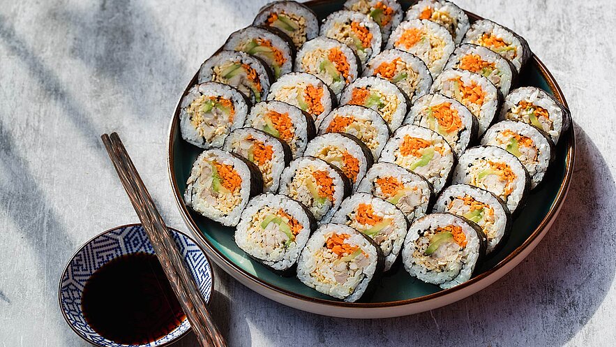 Ein Teller mit mehreren Sushi-Rollen, die mit Reis, Seealgen und bunten Füllungen wie Karotten und Gemüse gefüllt sind. Neben dem Teller liegen Essstäbchen und eine kleine Schüssel mit Sojasauce.