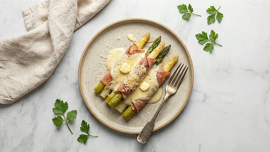 Ein Teller mit grünem und weißem Spargel, umwickelt mit Schinken, überzogen mit einer cremigen Sauce und Käse. Ein Stück Butter ist auf dem Gericht platziert. Neben dem Teller liegen ein Besteck und einige frische Petersilienblätter.
