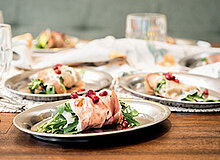 Auf einem Holztisch stehen mehrere silberne Teller mit frischen Salatgerichten. Jedes Gericht enthält Rucola, umwickelt mit Schinken und garniert mit Granatapfelkernen. Der Hintergrund zeigt verschwommene Gläser und weitere Teller.