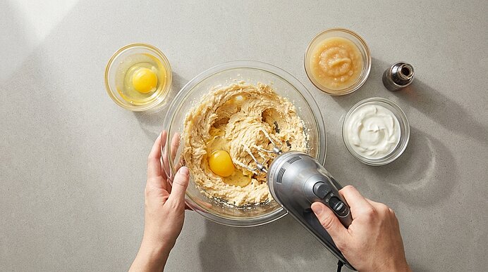Zwei Hände halten eine Glasschüssel und einen Handmixer, der gerade Teig mit Eiern verrührt; daneben kleine Schalen mit weiteren Zutaten.