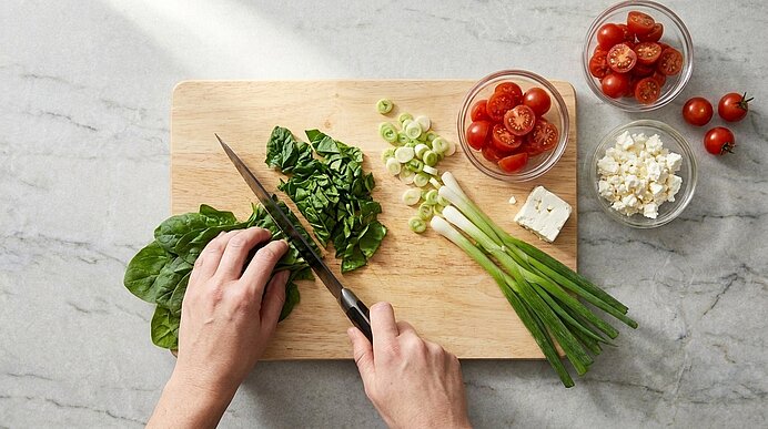 Hände schneiden Spinat mit einem Messer auf einem Holzbrett; daneben Frühlingszwiebeln, halbierte Cocktailtomaten in Schalen und Feta-Käse — Vorbereitung für einen Salat.