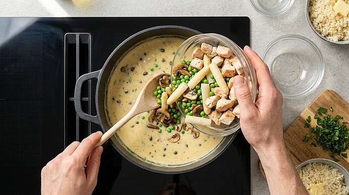 Hände geben gewürfeltes Hähnchen, grüne Erbsen und Spargel in einen Topf mit cremiger Pilzsauce, während auf dem Herd mit einem Holzlöffel gerührt wird.